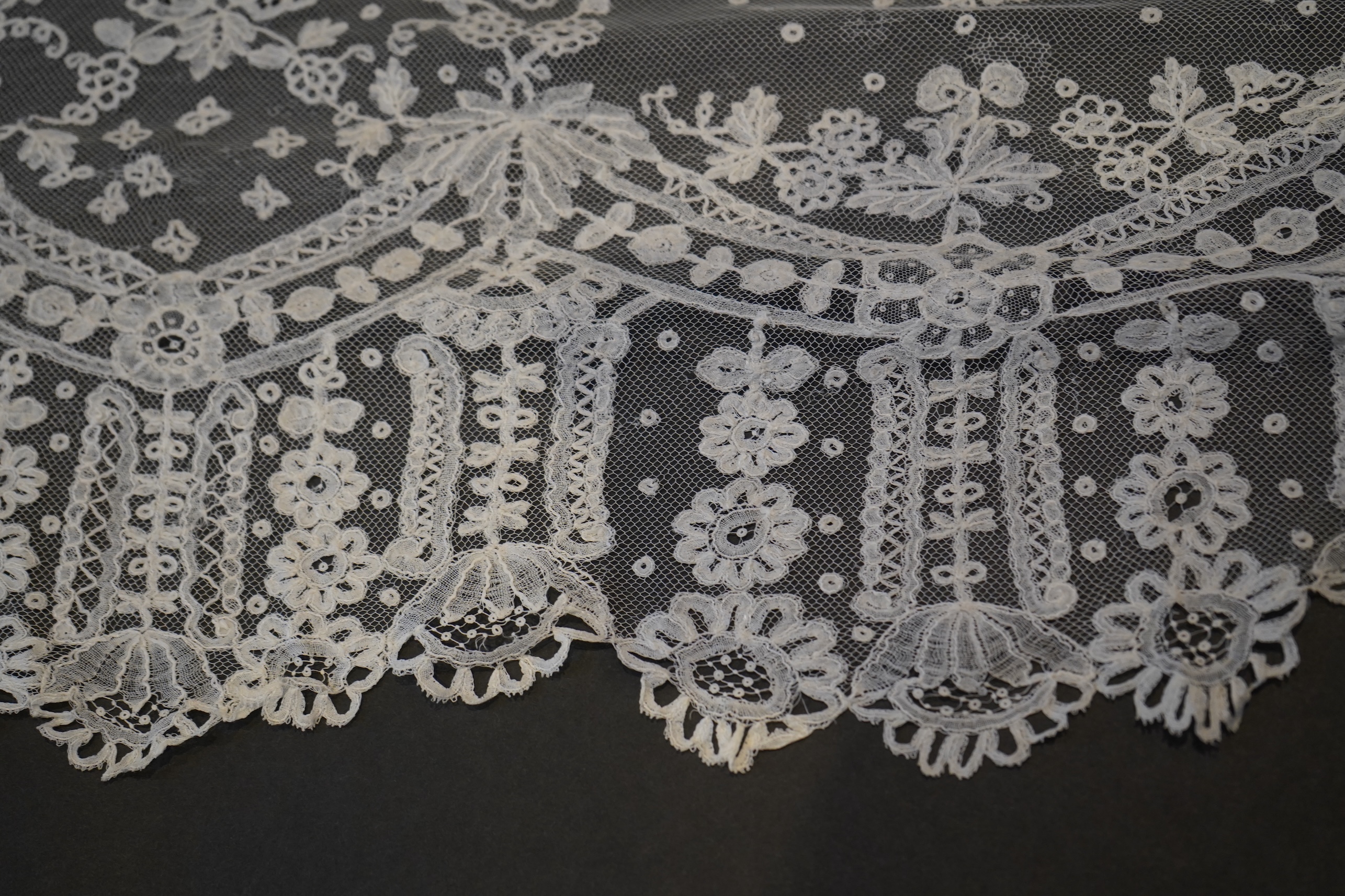 A deep elaborately designed flounce of mid 19th century Brussels bobbin appliquéd lace on net, with finished lace ends, worked with an intricate floral border below large floral cartouches framed in similar motifs and al